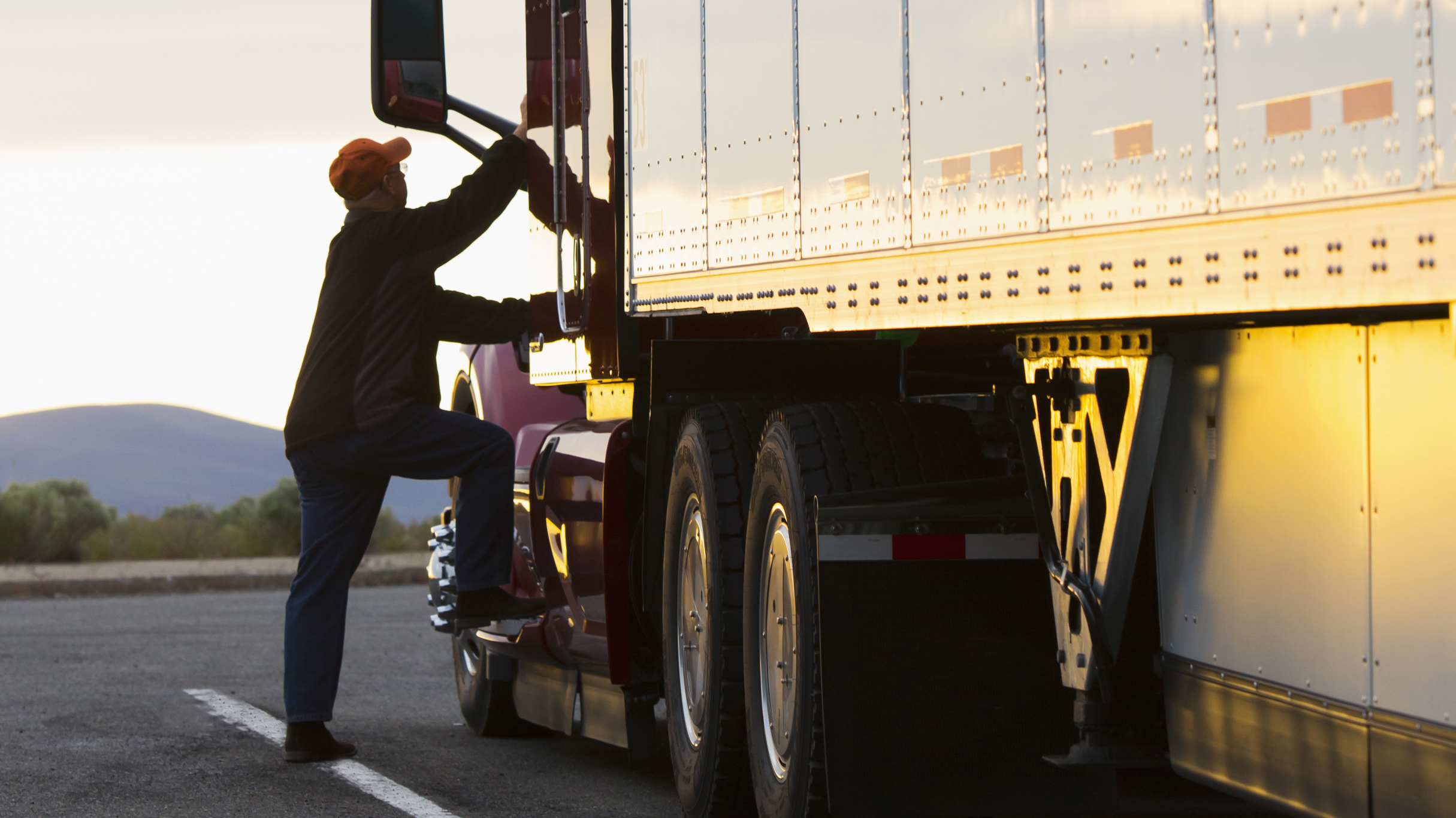 Commercial truck driver climbing into driver’s side of 18 wheeler semi-truck cab during sunset. 